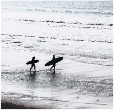 Two people walking along the beach, through ankle-deep surf, with their surfboards tucked under their arms.